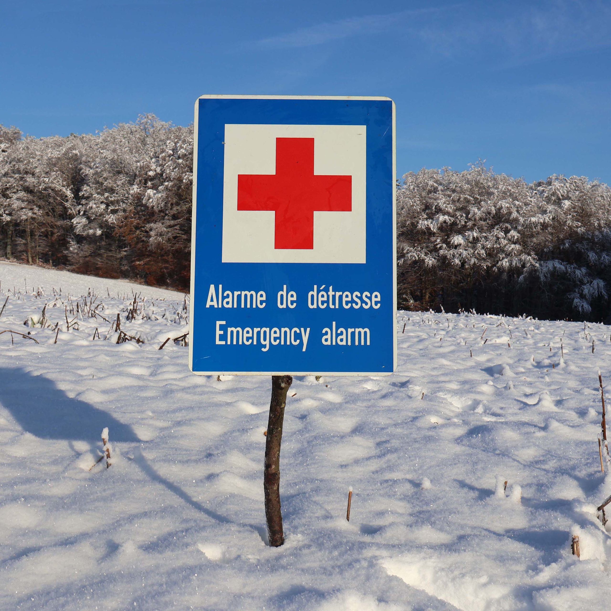 Panneau de signalisation Suisse, avec logo croix rouge, sur fond blanc. Avec bordure bleue. Texte alarme de détresse et en Anglais : Emergency alarm